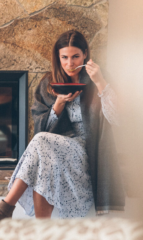 Weiblicher Gast sitz an kalten Tagen vor dem Kamin und wärmt sich mit einer Decke und einer Suppe aus der Küche des Hotel.
