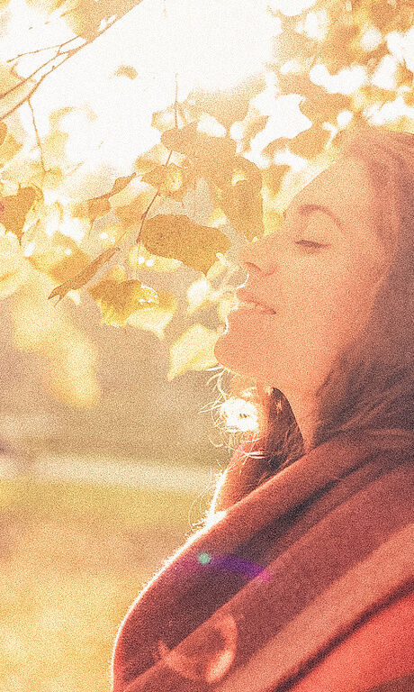 Eine Frau genießt die wärmende Sonne im Gesicht unter einem herbstlichen Blätterdach.