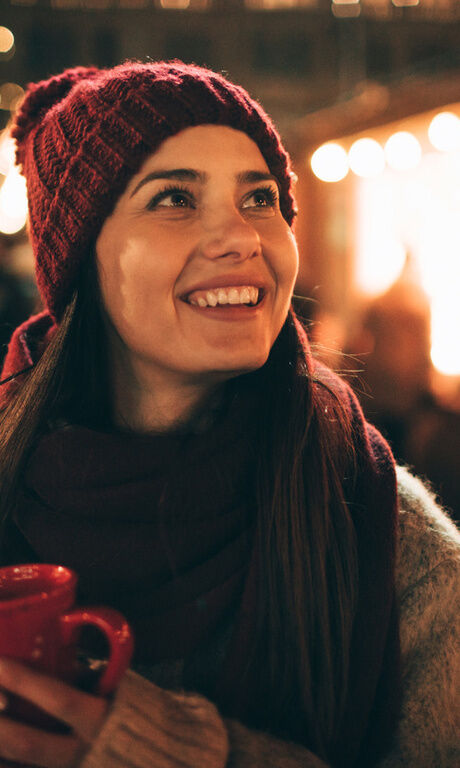 Frau erfreut sich an Lichtern und der Stimmung des Weihnachtsmarkts. Warm angezogen hält sie ein heißes Getränk in der Hand.