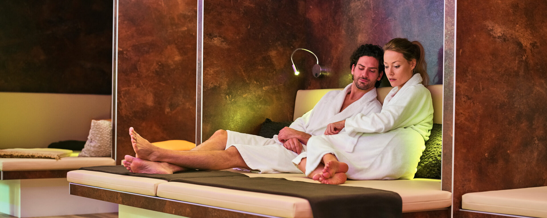 Ein Paar entspannt sich auf einem Bett im Ruheraum des Wellnessbereichs im Hotel Eibl-Brunner. Eingehüllt in flauschige Bademäntel.